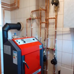 Impact Jakub Bauer - Czerwono-szary piec ATMOS S Genrator z czarnym kominem i miedzianymi rurami instalacji centralnego ogrzewania na tle kafelkowanej ściany.