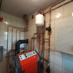 Impact Jakub Bauer - Czerwony piec grzewczy z czarnym kominem i licznymi miedzianymi rurami instalacji grzewczej na tle kafelkowanej ściany.