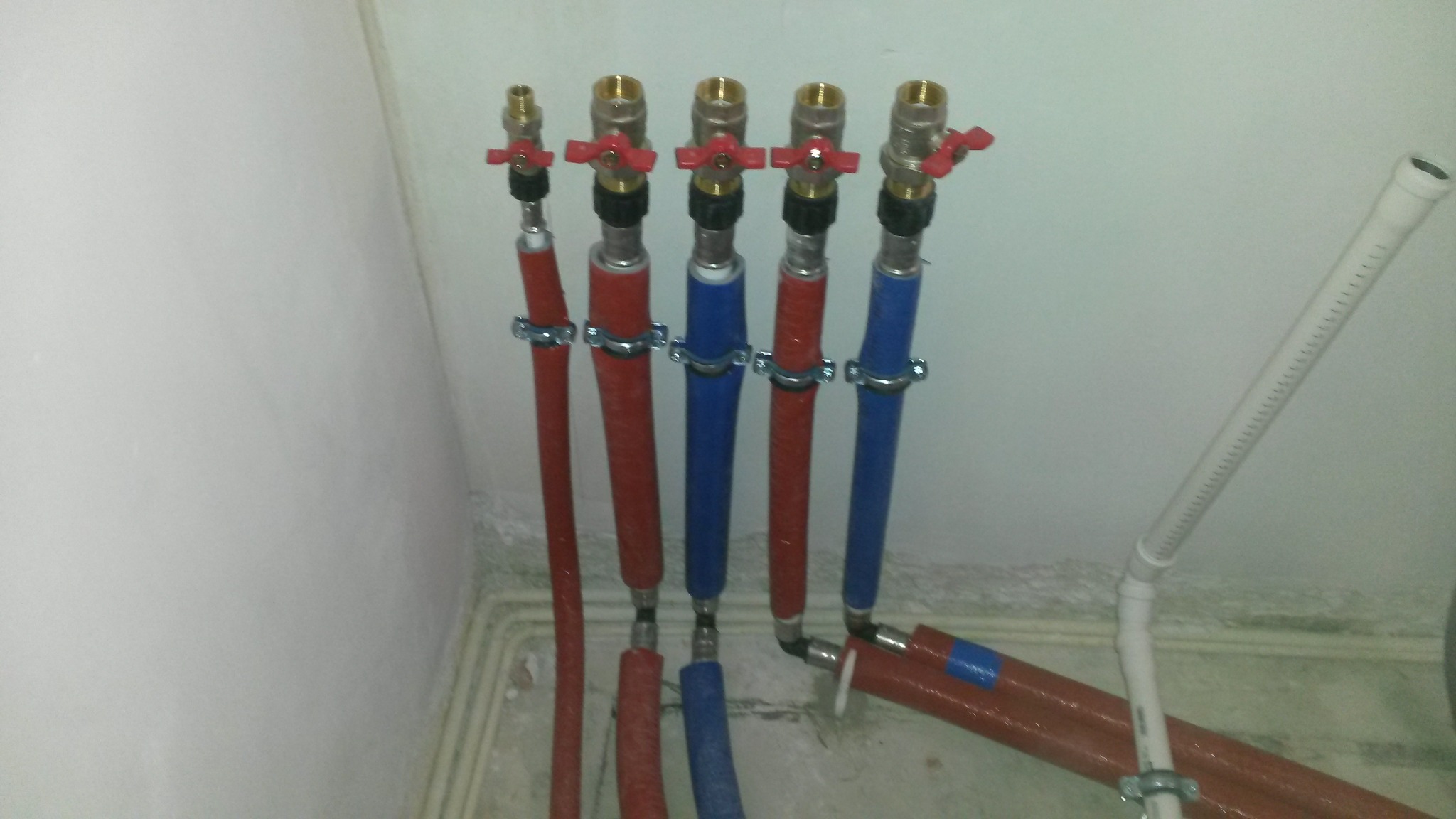 Różnokolorowe rury instalacji wodnej (czerwone i niebieskie) z zaworami, przymocowane do ściany za pomocą metalowych uchwytów, widoczne łączenia i fragmenty innych rur.