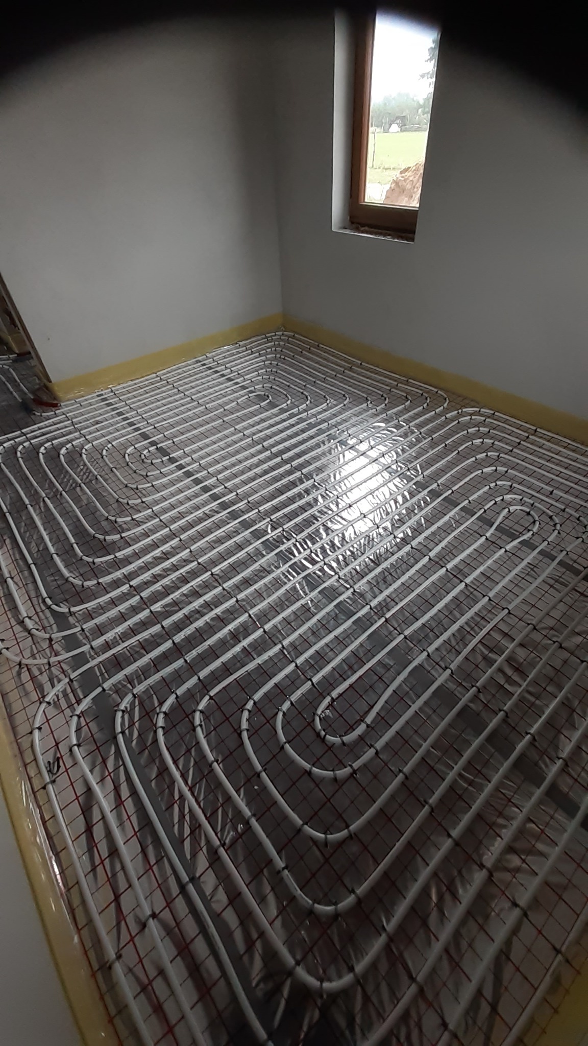 Instalacja ogrzewania podłogowego z białych rur na folii izolacyjnej w jasnym pomieszczeniu z oknem.