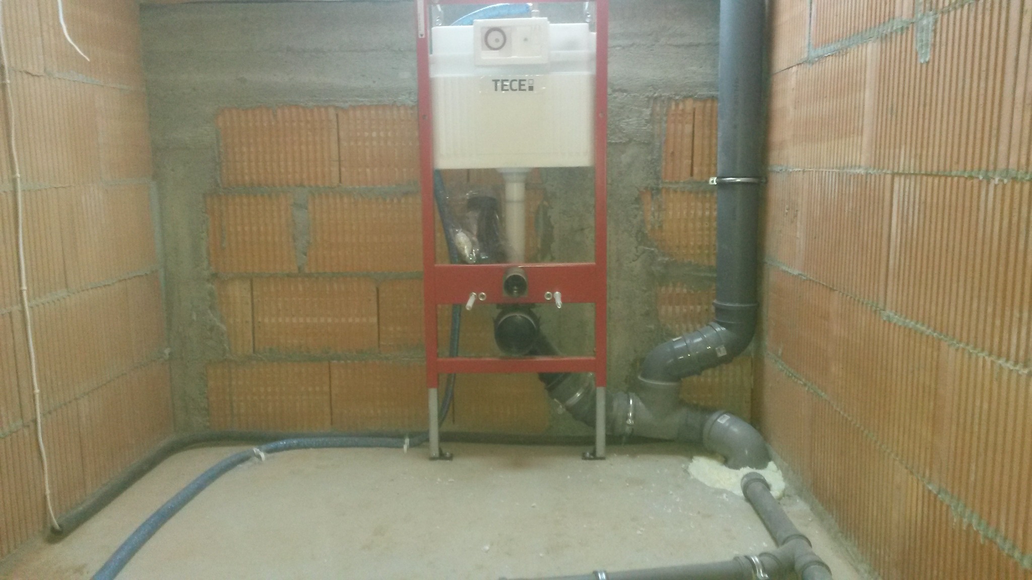 Stelaż podtynkowy TECE zamontowany w surowym pomieszczeniu z cegły, widoczne rury kanalizacyjne i dopływ wody.