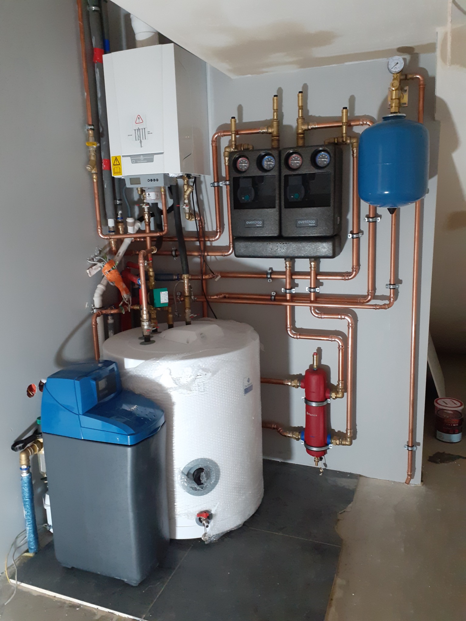 Kompleksowa instalacja grzewcza w kotłowni: biały zasobnik ciepłej wody owinięty folią, szary zmiękczacz wody DWT, czarny moduł hydrauliczny Oventrop, niebieskie naczynie przeponowe oraz piec...