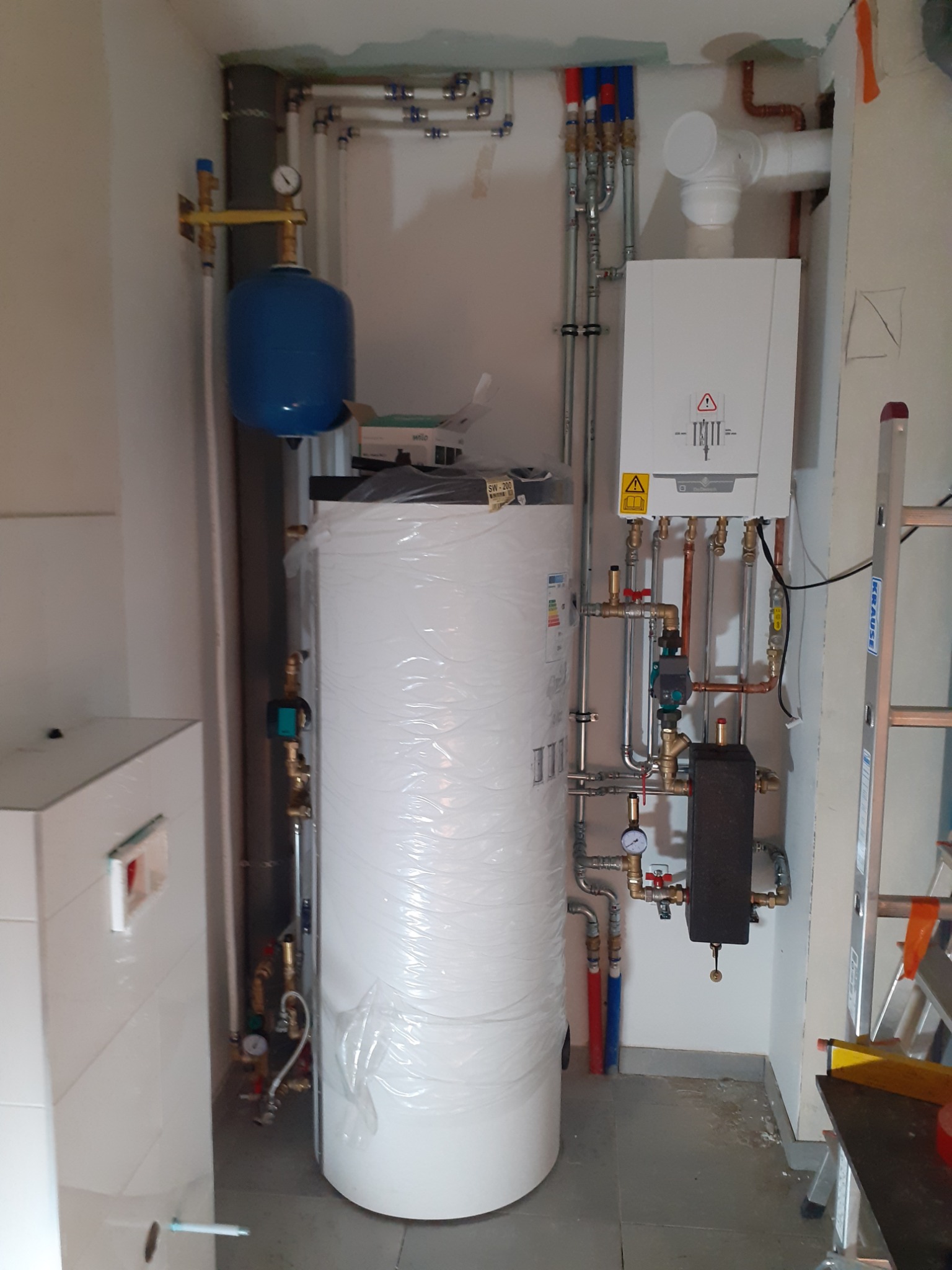 Kotłownia w trakcie montażu: biały zasobnik ciepłej wody owinięty folią, piec gazowy, rury miedziane i stalowe, manometry, drabina oparta o ścianę.