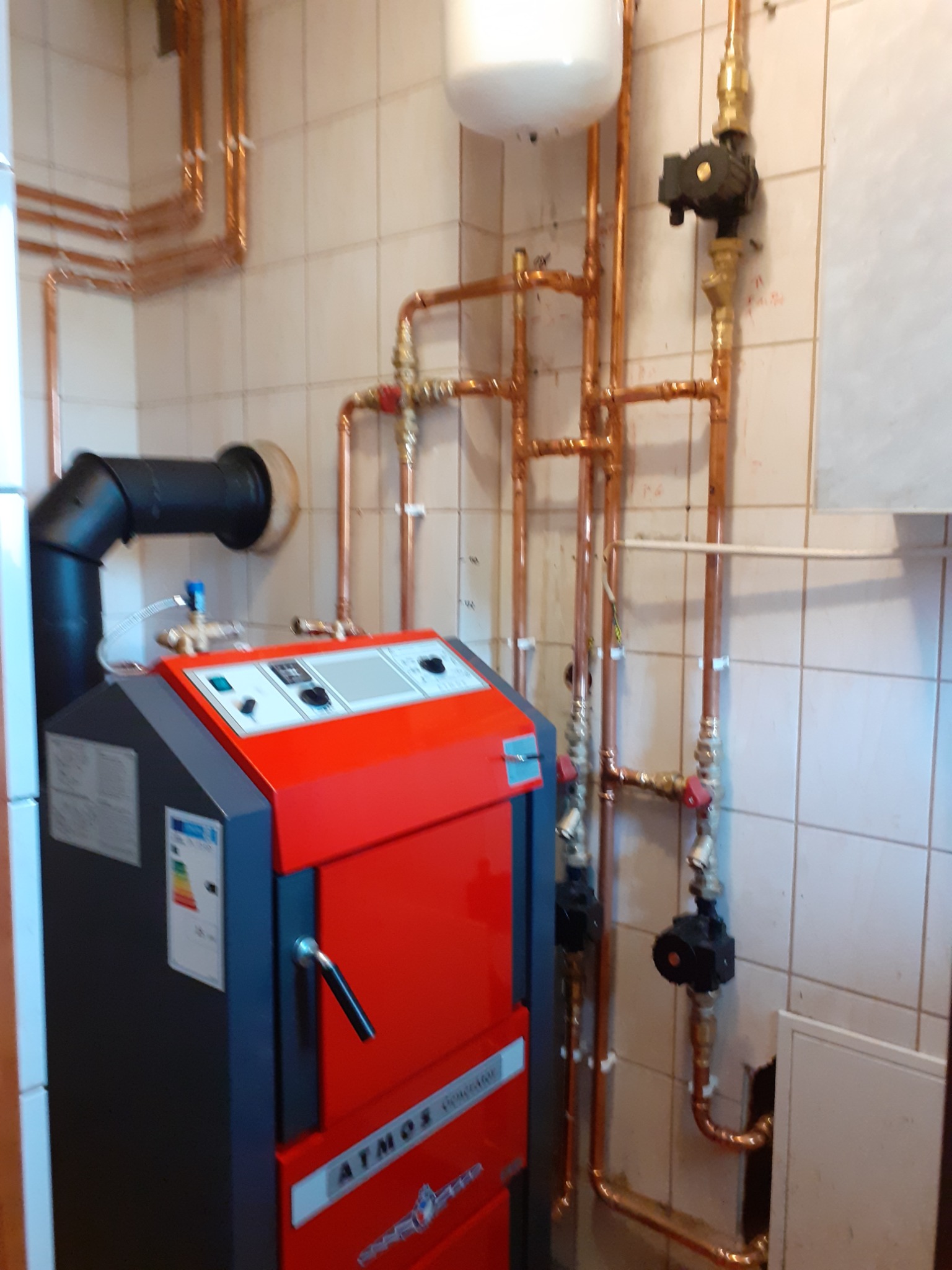 Czerwono-szary piec ATMOS S Genrator z czarnym kominem i miedzianymi rurami instalacji centralnego ogrzewania na tle kafelkowanej ściany.