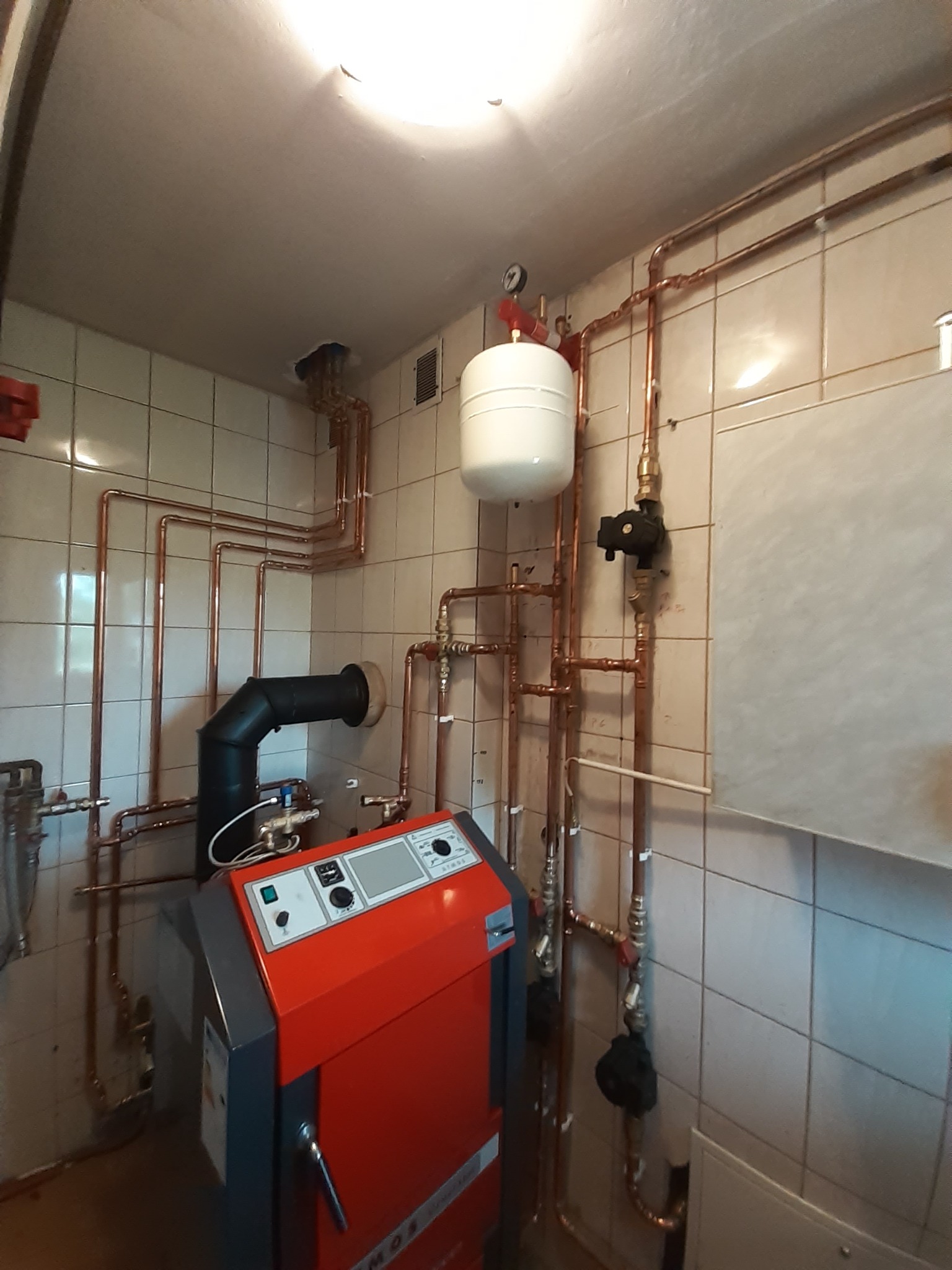 Czerwony piec grzewczy z czarnym kominem i licznymi miedzianymi rurami instalacji grzewczej na tle kafelkowanej ściany.