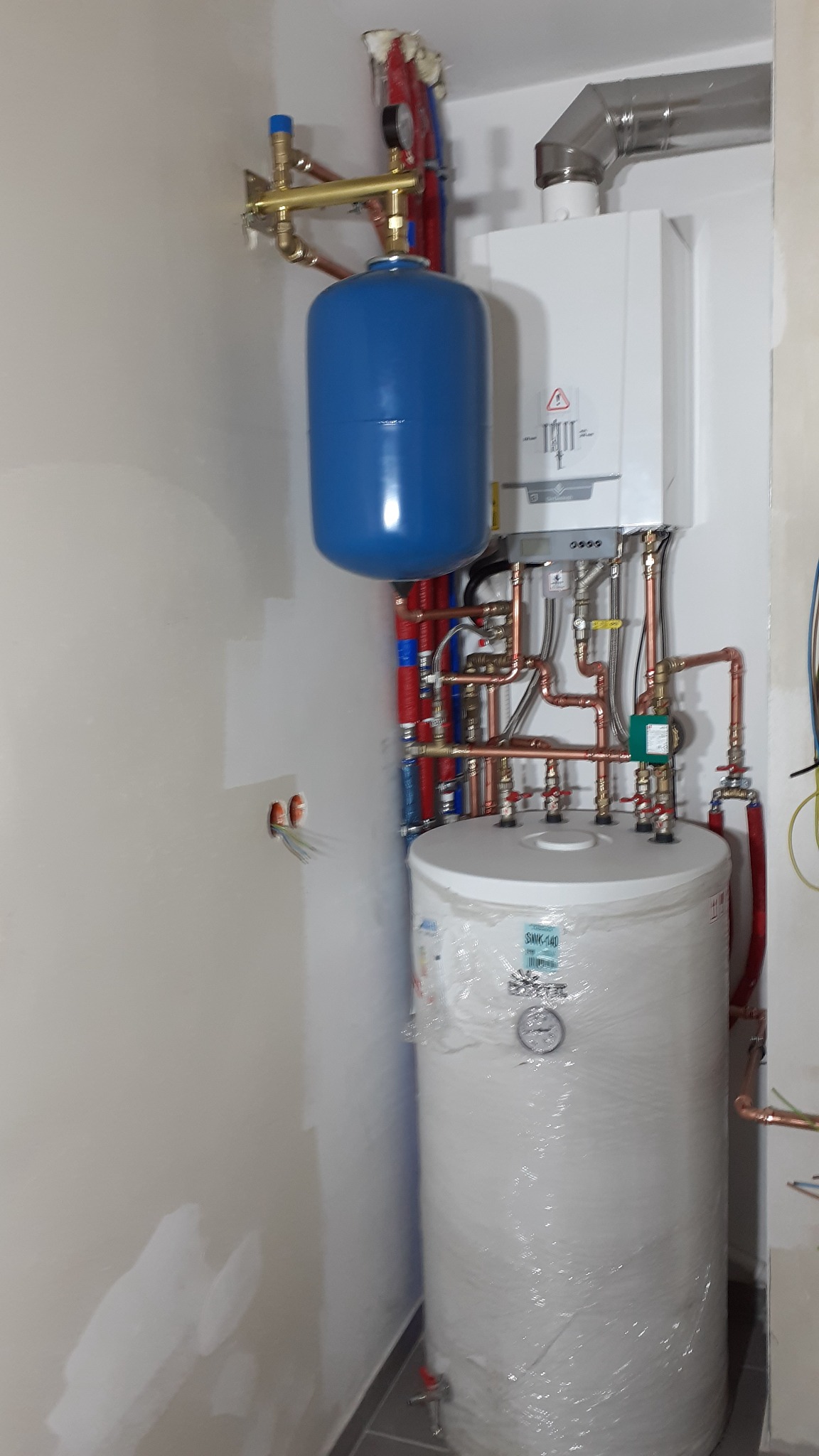 Nowa instalacja centralnego ogrzewania z piecem gazowym, zbiornikiem buforowym i licznymi miedzianymi rurami w surowym wnętrzu z widocznymi śladami po malowaniu na ścianie.