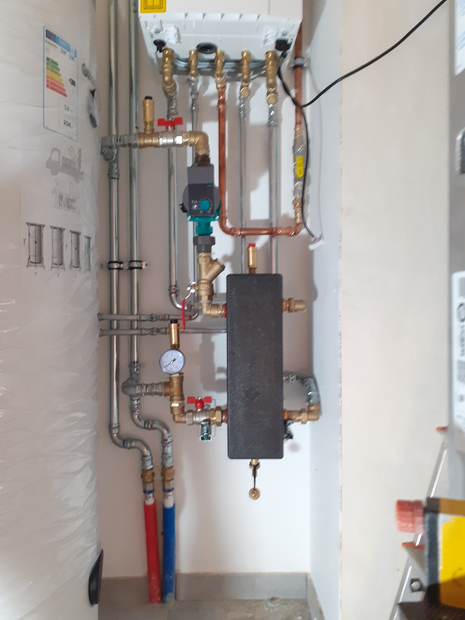Węzeł cieplny z czarnym wymiennikiem ciepła, manometrem i zaworami, widoczne rury miedziane i stalowe, instalacja w pomieszczeniu technicznym, na ścianie naklejka z klasą energetyczną urządzenia.