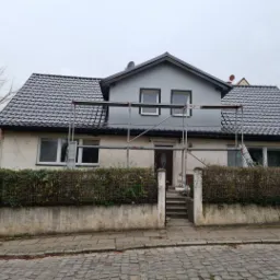 Dom jednorodzinny z szarą elewacją i nowym, błyszczącym, ciemnym dachem, otoczony rusztowaniem, widok z poziomu ulicy.