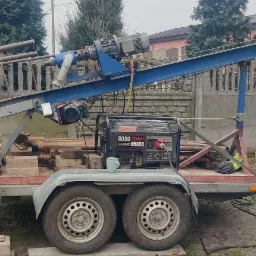 Mobilny zestaw wiertniczy na przyczepie z generatorem prądu PEZAL 8000, rurami wiertniczymi i kubkiem na kole, zaparkowany obok samochodu z napisem 'studnie głębinowe'.