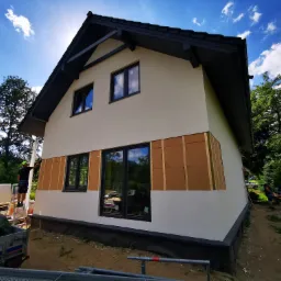 Montaż płyt izolacyjnych na elewacji domu jednorodzinnego, widoczne rusztowanie, pracownik na drabinie i częściowo ocieplona ściana z czarnymi ramami okiennymi.