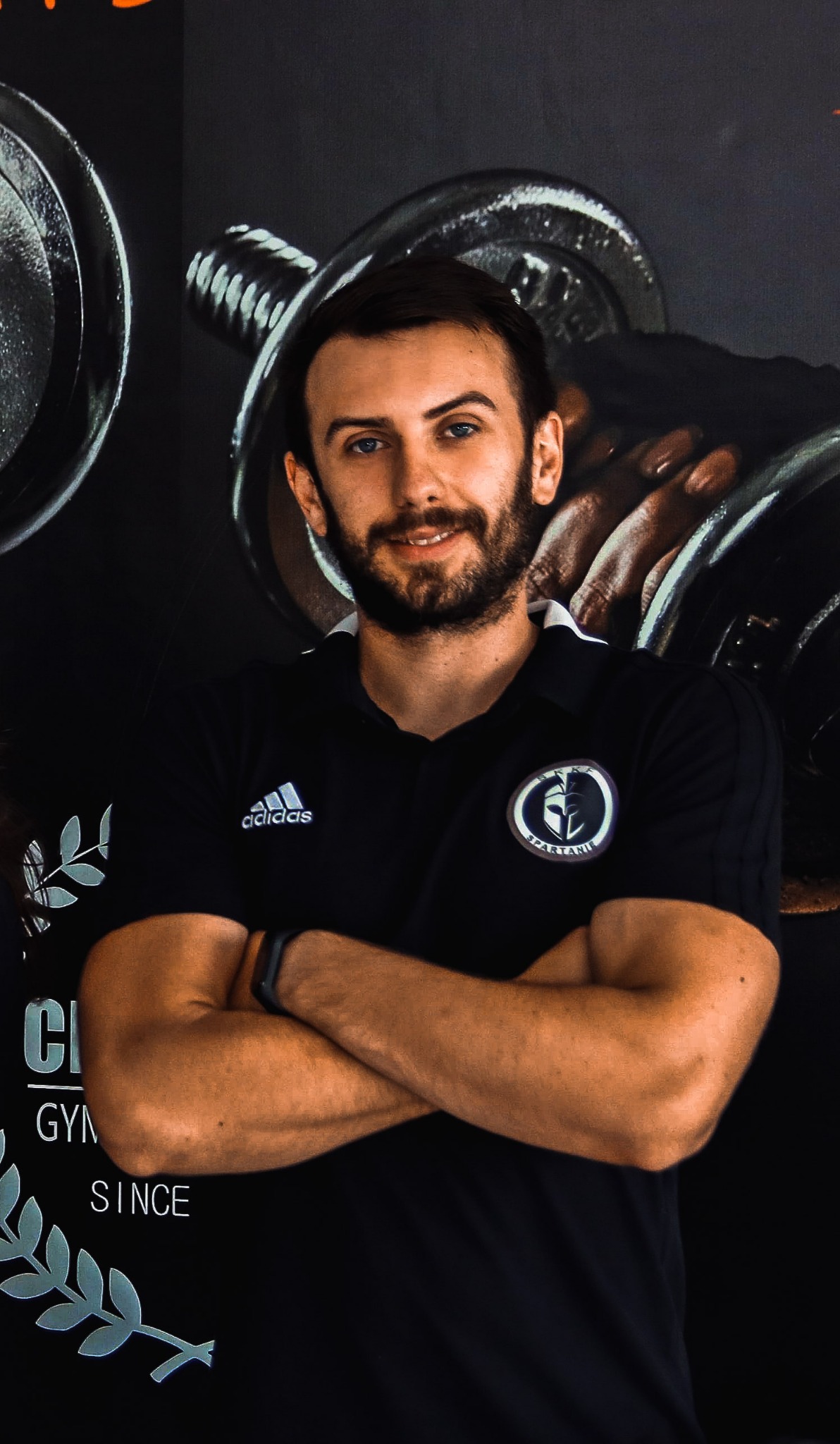 Młody mężczyzna z brodą, ubrany w czarną koszulkę sportową z logo Adidas i emblematem klubu sportowego, stoi na tle plakatu z ciężarami i napisem 'Classic Gym Since'.