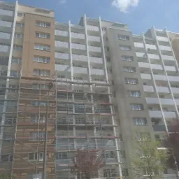 Wielopiętrowy budynek mieszkalny w trakcie prac remontowych elewacji, z widocznymi rusztowaniami obejmującymi część fasady, oraz fragmenty odnowionej i surowej powierzchni. Ujęcie z dołu,...