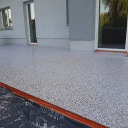 Wykończony taras z żywicy epoksydowej w odcieniach szarości i czerwieni, widoczne zabezpieczenia pomarańczową taśmą przy ścianie budynku i drzwiach tarasowych, częściowo zakryty czarną folią ochronną.