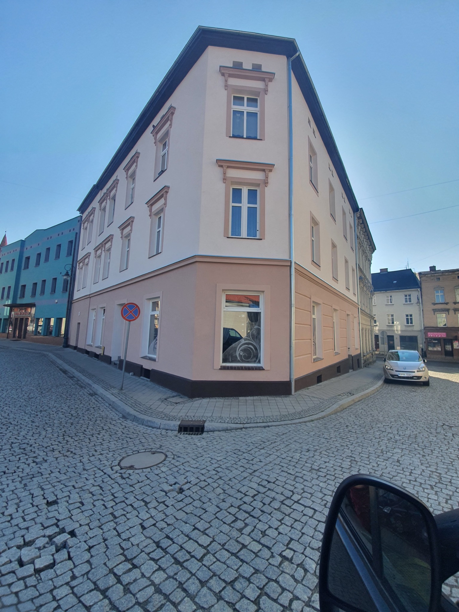 Odnowiona kamienica narożna w pastelowych kolorach z dekoracyjnymi elementami architektonicznymi, widok z perspektywy ulicy wyłożonej kostką brukową w słoneczny dzień.