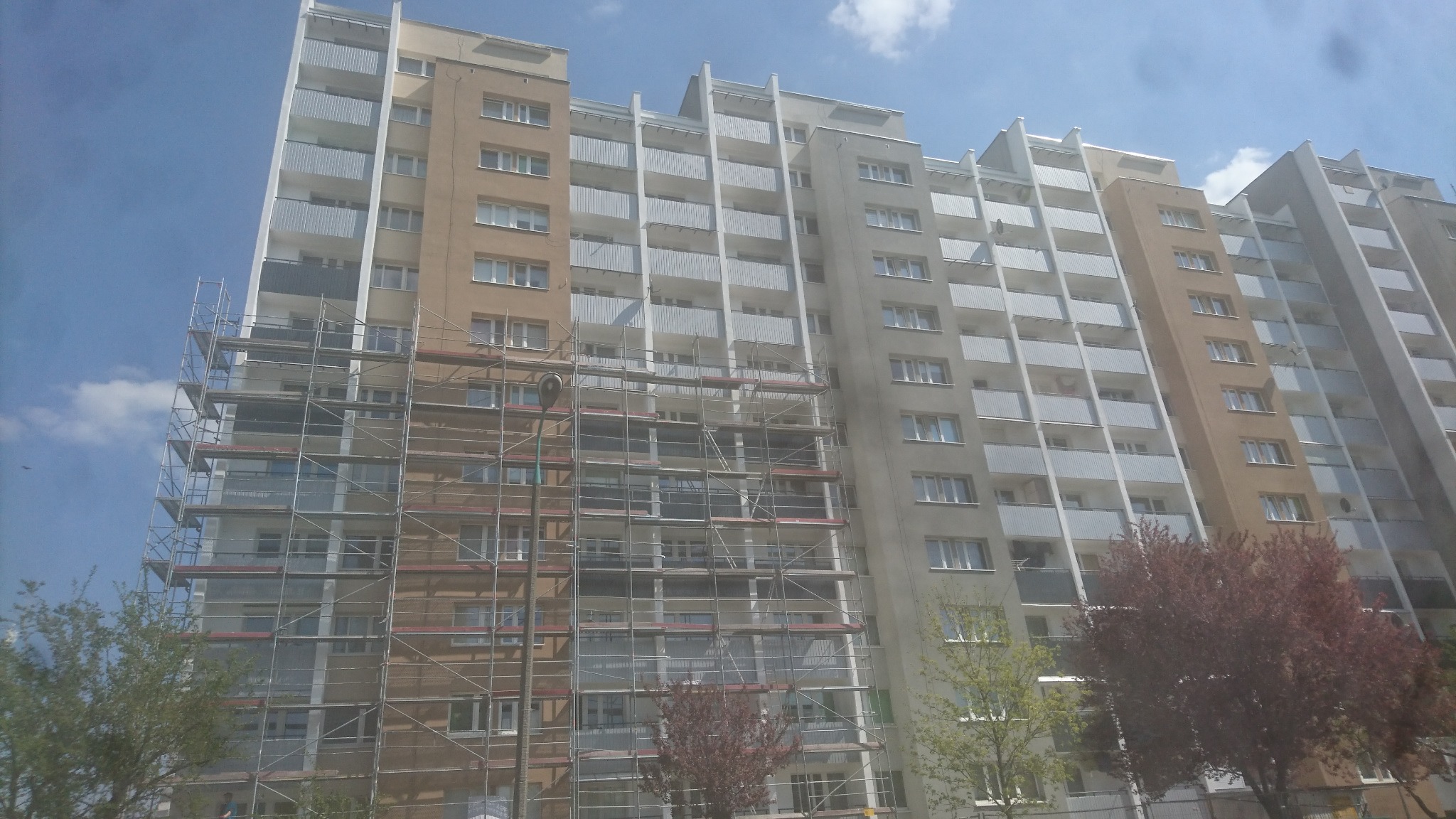 Wielopiętrowy budynek mieszkalny w trakcie prac remontowych elewacji, z widocznymi rusztowaniami obejmującymi część fasady, oraz fragmenty odnowionej i surowej powierzchni. Ujęcie z dołu,...