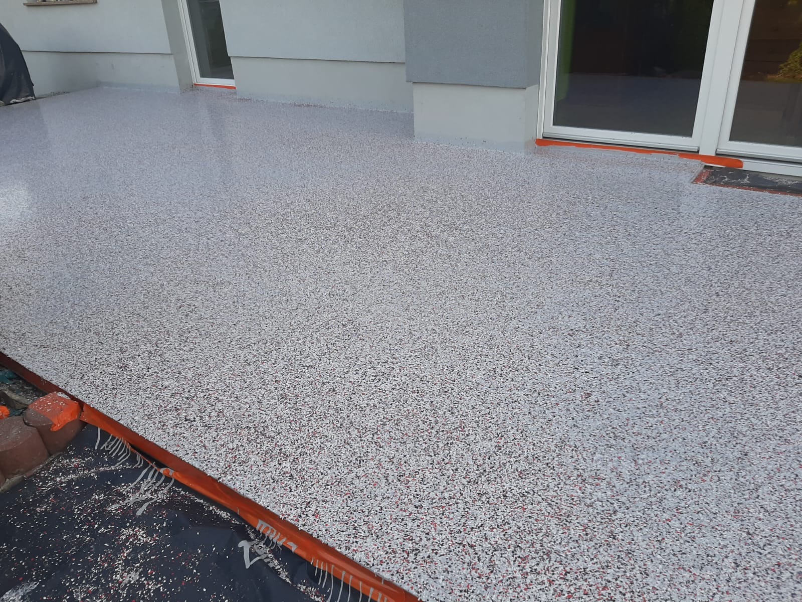 Nowa, jasnoszara posadzka żywiczna z drobnymi, kolorowymi granulkami na tarasie domu jednorodzinnego, widoczne zabezpieczenia pomarańczową folią malarską przy oknach przesuwnych i krawędzi tarasu.