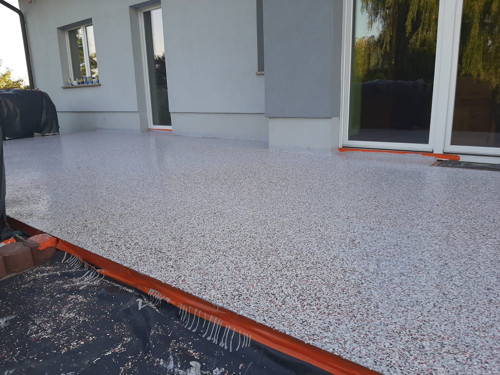 Wykończony taras z żywicy epoksydowej w odcieniach szarości i czerwieni, widoczne zabezpieczenia pomarańczową taśmą przy ścianie budynku i drzwiach tarasowych, częściowo zakryty czarną folią ochronną.