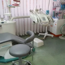 Wnętrze gabinetu z fotelem dentystycznym, unit stomatologiczny z licznymi końcówkami, model szczęki na półce, reklamy pasty do zębów Meridol na ścianie i wentylator pod nimi.