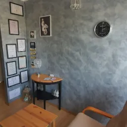 Poczekalnia: ściana z dekoracyjnym tynkiem w odcieniach szarości, zegar ścienny, stolik z lampką, krzesło z drewnianymi podłokietnikami, drzwi z naklejkami informacyjnymi i certyfikaty na ścianie.