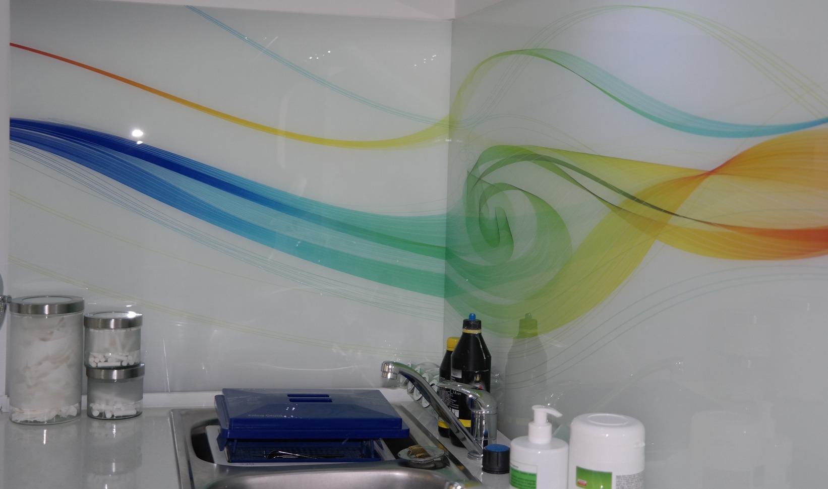 Biała ściana w gabinecie z abstrakcyjnym wzorem w kolorach niebieskim, zielonym, żółtym i pomarańczowym, widoczny zlew ze srebrną baterią, pojemniki na waciki i płyny do dezynfekcji.