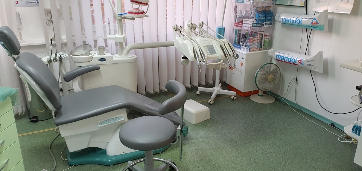 Wnętrze gabinetu z fotelem dentystycznym, unit stomatologiczny z licznymi końcówkami, model szczęki na półce, reklamy pasty do zębów Meridol na ścianie i wentylator pod nimi.