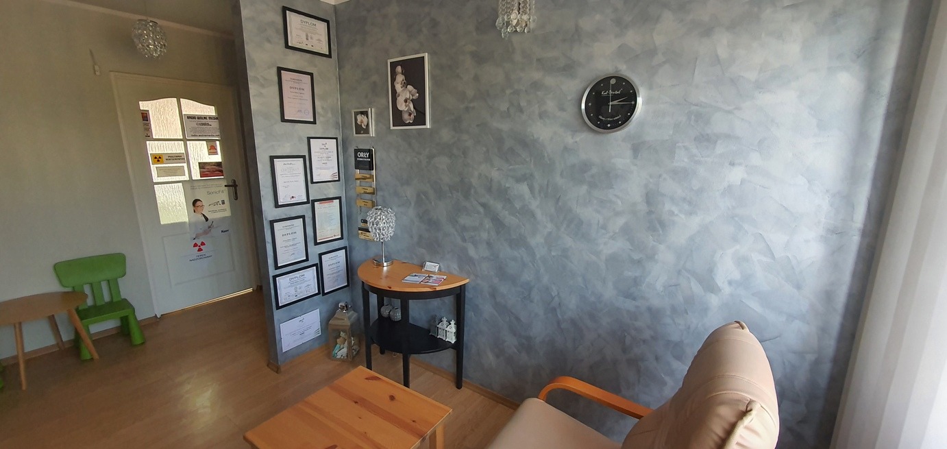 Poczekalnia: ściana z dekoracyjnym tynkiem w odcieniach szarości, zegar ścienny, stolik z lampką, krzesło z drewnianymi podłokietnikami, drzwi z naklejkami informacyjnymi i certyfikaty na ścianie.