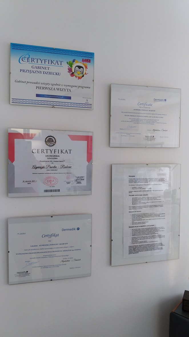 Ściana gabinetu z powieszonymi certyfikatami i dyplomami, w tym certyfikat 'Gabinet Przyjazny Dziecku' z logo z pingwinem oraz certyfikaty Dermedik