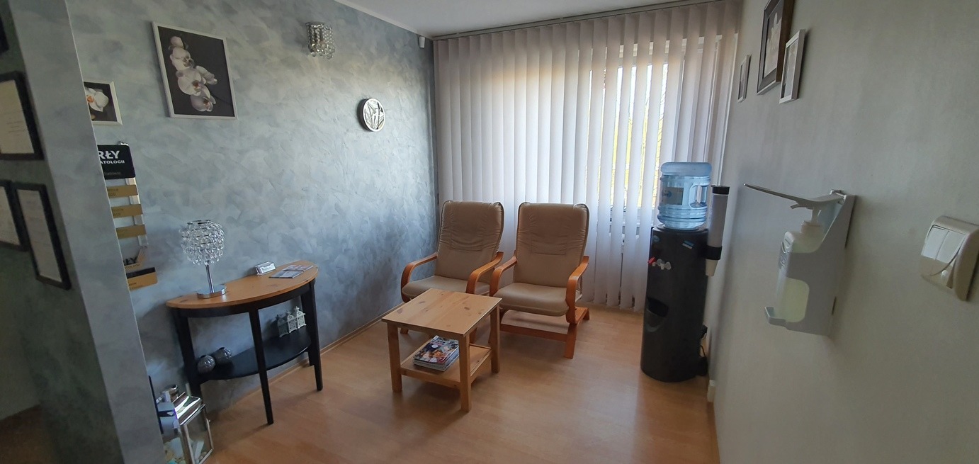 Poczekalnia z dwoma fotelami, stolikiem kawowym, dystrybutorem wody i dozownikiem do dezynfekcji rąk w gabinecie lekarskim, ściany w odcieniach szarości, na ścianach wiszą obrazy i zegar.