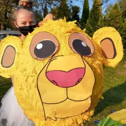 Dziewczyna w masce trzyma dużą, żółtą piniatę w kształcie głowy lwa, z wyraźnymi uszami i czarnymi oczami, na tle zieleni i błękitnego nieba.