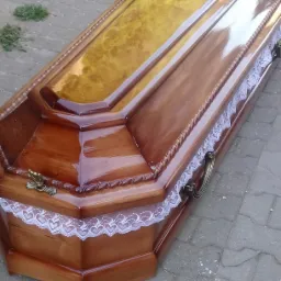 Lakierowana drewniana trumna z krzyżem i koronkowym wykończeniem, sfotografowana z perspektywy bocznej na brukowanej powierzchni.