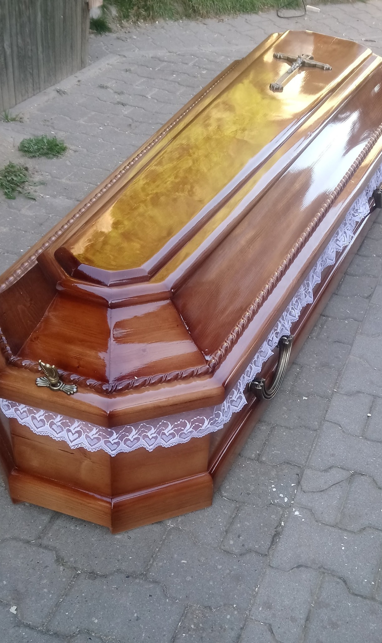 Lakierowana drewniana trumna z krzyżem i koronkowym wykończeniem, sfotografowana z perspektywy bocznej na brukowanej powierzchni.