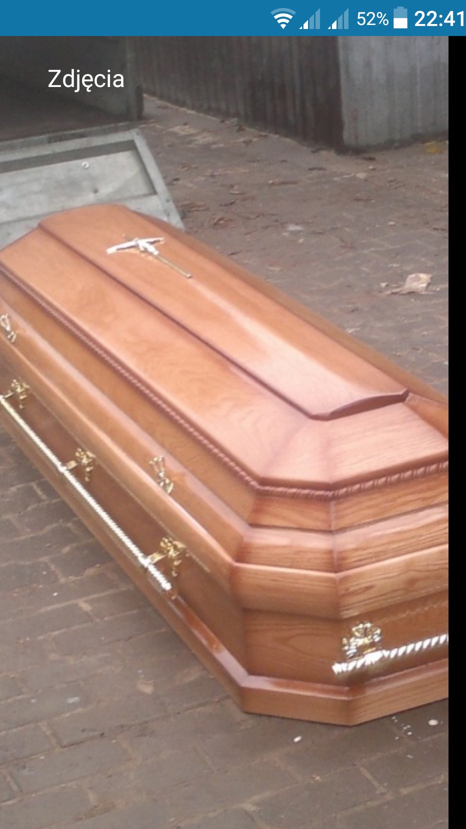 Lakierowana, dębowa trumna z metalowymi uchwytami i krzyżem na wieku, sfotografowana na zewnątrz na brukowanej powierzchni.