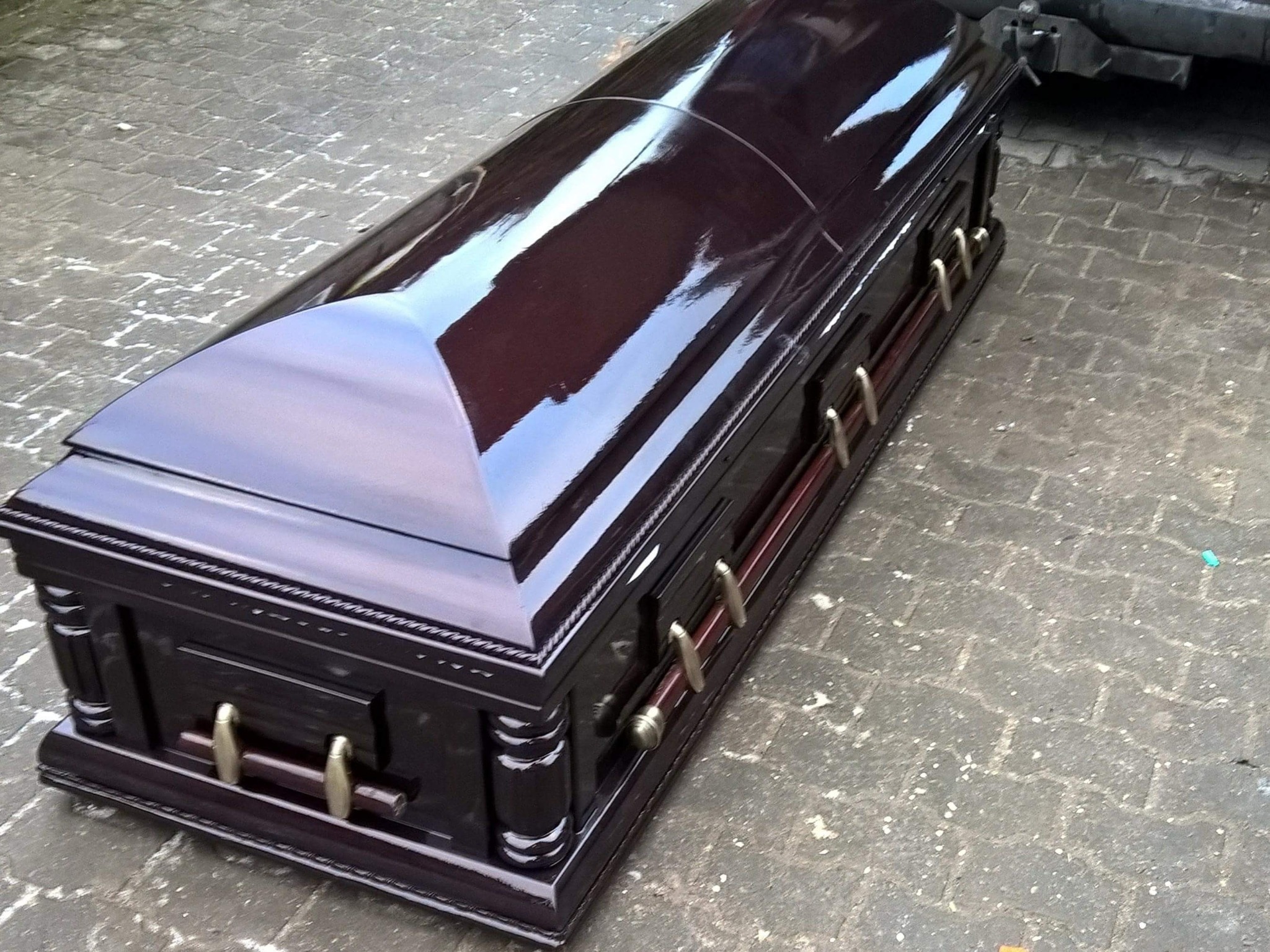 Lakierowana, mahoniowa trumna z metalowymi uchwytami na szarym brukowanym podłożu, widoczny fragment pojazdu w tle.