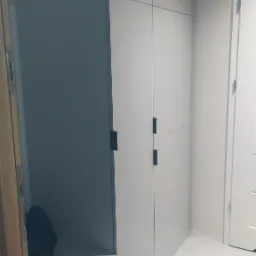 Biała szafa wnękowa z czarnymi, prostokątnymi uchwytami, z otwartą półką na buty i kurtki, na jasnej podłodze, widok z perspektywy podłogi.