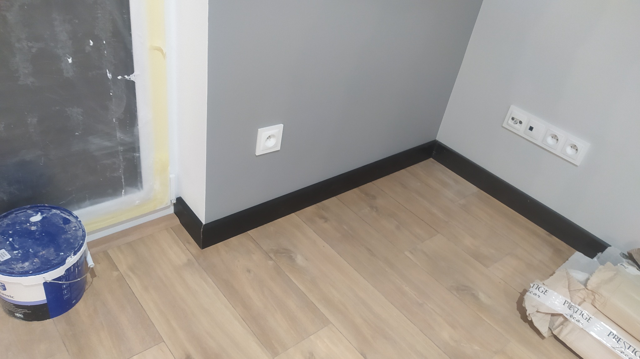 Przygotowanie do malowania: wiadro z farbą przy osłoniętej taśmą ścianie, pomalowana szara ściana z gniazdkiem, ułożone panele podłogowe i listwy przypodłogowe.