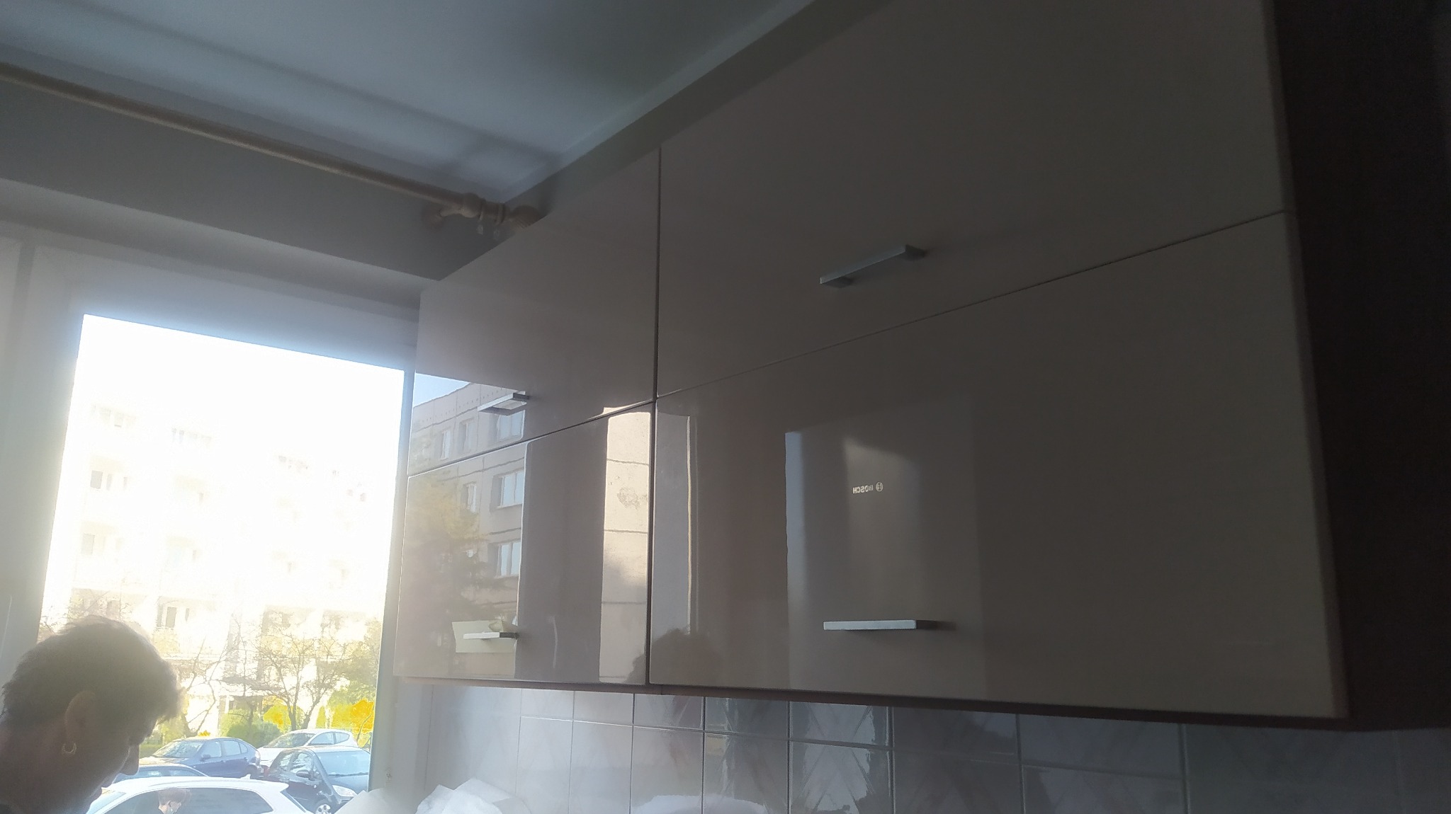 Beżowe, lakierowane szafki kuchenne wiszące, z prostymi, metalowymi uchwytami, odbijające światło okna i zabudowę zewnętrzną.