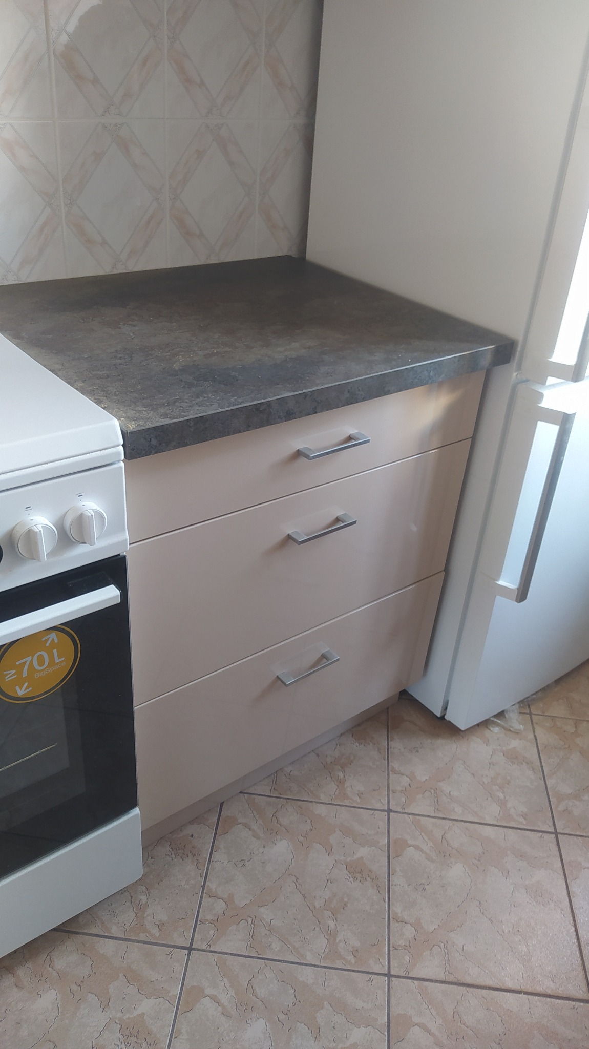 Fragment kuchni z beżowymi szafkami z trzema szufladami z metalowymi uchwytami, obok biała kuchenka i lodówka, na podłodze płytki w odcieniach beżu i brązu, na ścianie płytki w romby.