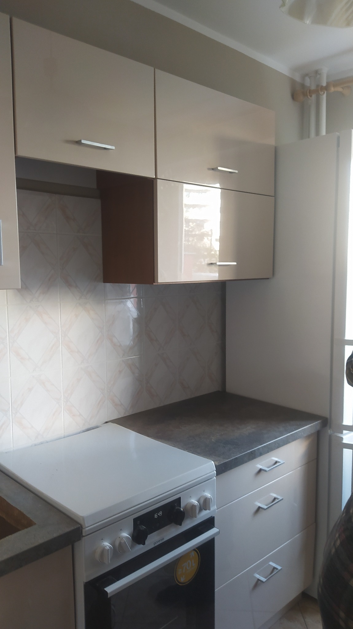 Jasna kuchnia z błyszczącymi frontami szafek górnych i dolnych, biała kuchenka z czarnym piekarnikiem, płytki ścienne w romby, blat w ciemnym kolorze.