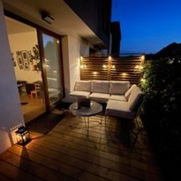 MARCUS Usługi Remontowe Marek Frydrychowski - Oświetlony balkon z drewnianą podłogą i jasną sofą narożną. Lampion na podłodze, dekoracyjne lampki na drewnianej ściance. Widok na nocny krajobraz.