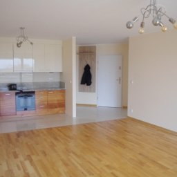 MARCUS Usługi Remontowe Marek Frydrychowski - Jasne wnętrze mieszkania z drewnianą podłogą, zabudową kuchenną i nowoczesnym oświetleniem. Widoczny fragment przedpokoju z wieszakiem i wejściem.