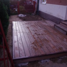 MARCUS Usługi Remontowe Marek Frydrychowski - Nowy taras drewniany z niskimi schodkami w Sławoszynie, wykonany z desek kompozytowych. Widoczne ślady wilgoci na powierzchni, w tle siatka ogrodzeniowa.
