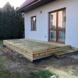 MARCUS Usługi Remontowe Marek Frydrychowski - Nowy, drewniany taras z niskimi schodami przy białym domu, otoczony zielenią. Widoczne detale konstrukcji i naturalna faktura drewna.