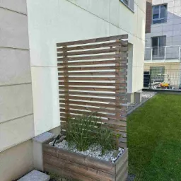 Nowoczesna drewniana ścianka działowa z poziomych listew, połączona z podwyższoną rabatą, wypełniona roślinami i białym żwirem przy budynku.