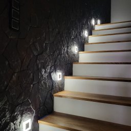 MARCUS Usługi Remontowe Marek Frydrychowski - Schody z drewnianymi stopniami i oświetleniem LED wbudowanym w ciemną, kamienną ścianę. Nowoczesne wykończenie wnętrza.
