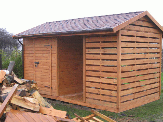 Drewniana altana z zamykaną częścią i ażurową ścianą boczną, brązowy dach z gontu bitumicznego, sterta drewna obok. Widok z zewnątrz w pochmurny dzień.