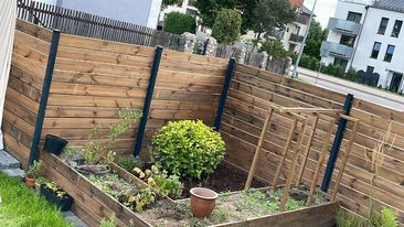 Drewniana konstrukcja w ogrodzie z rabatami i roślinami. Elementy drewniane w odcieniach brązu, tło stanowią budynki mieszkalne i zieleń.