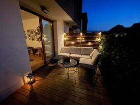 Oświetlony balkon z drewnianą podłogą i jasną sofą narożną. Lampion na podłodze, dekoracyjne lampki na drewnianej ściance. Widok na nocny krajobraz.