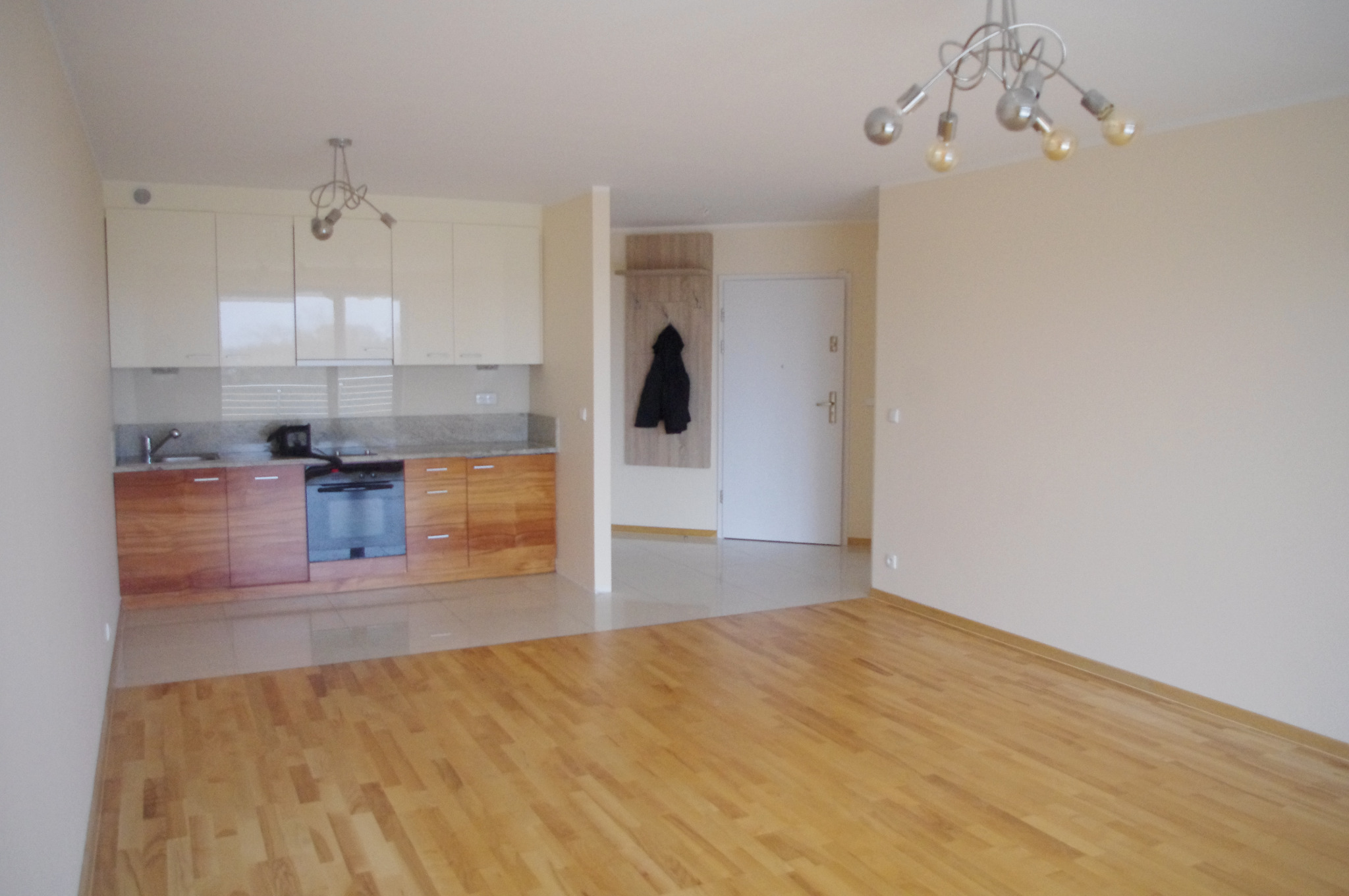 Jasne wnętrze mieszkania z drewnianą podłogą, zabudową kuchenną i nowoczesnym oświetleniem. Widoczny fragment przedpokoju z wieszakiem i wejściem.