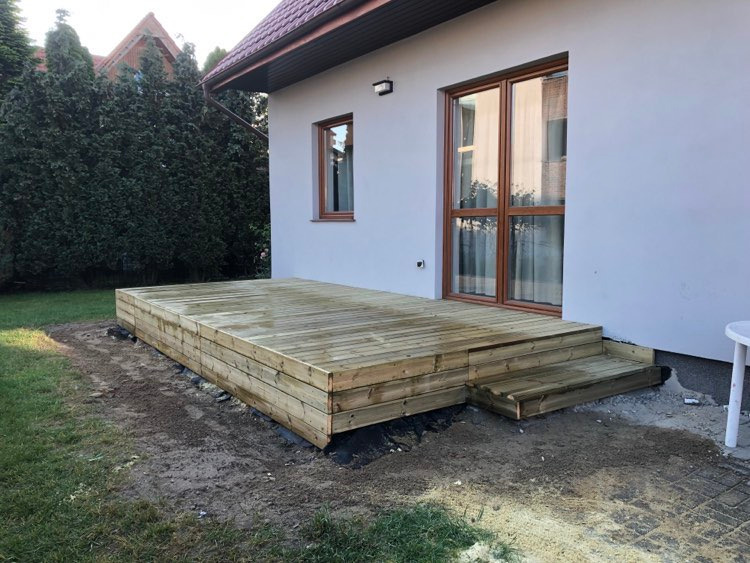 Nowy, drewniany taras z niskimi schodami przy białym domu, otoczony zielenią. Widoczne detale konstrukcji i naturalna faktura drewna.