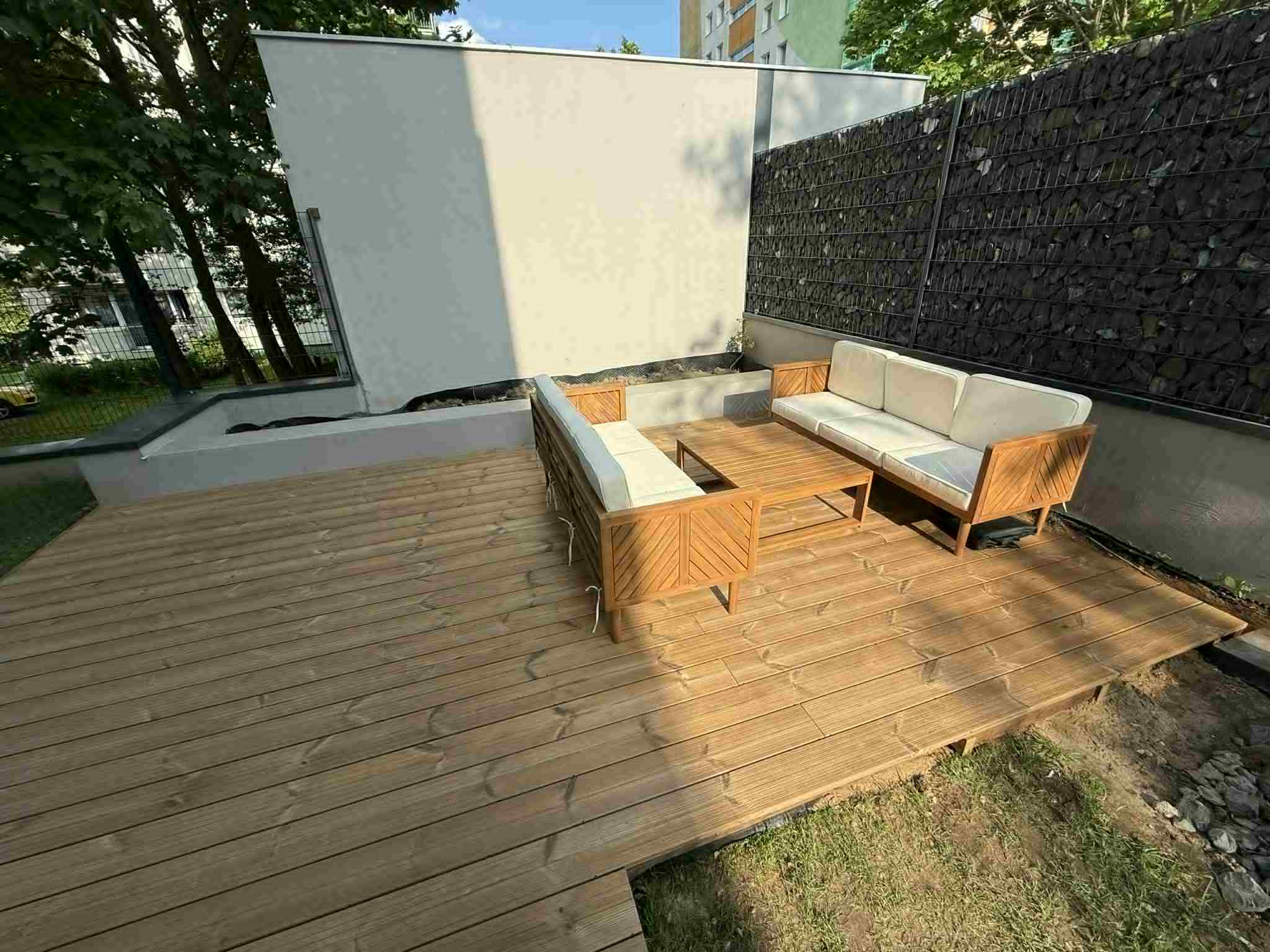 Jasny taras z drewna z nowoczesnym zestawem mebli ogrodowych i gabionami w tle. Widok z góry, słoneczny dzień.